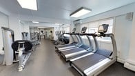 This single-floor fitness center offers a comprehensive workout environment designed for functionality and comfort. The space is outfitted with a prominent row of True-brand treadmills, each equipped with individual digital display consoles, lined along windowed walls that allow natural light to filter through roller shades. Alongside the treadmills, a series of elliptical machines provides additional cardio options. Toward the rear of the space, a dedicated strength-training area houses cable machines and various resistance equipment, offering a well-rounded selection for full-body workouts. Full-length mirrors along one wall enhance the sense of openness and allow users to monitor their form during exercise. The flooring consists of durable, dark rubberized material suited for high-traffic gym use. Overhead, large flush-mounted light panels supplement the natural light, ensuring the space remains well-illuminated throughout. A wall-mounted air conditioning unit contributes to climate control and comfort. The overall aesthetic is clean and minimalist, with white walls and a practical layout that maximizes usable floor space while maintaining clear circulation paths between equipment.