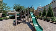 This outdoor playground offers a thoughtfully designed recreational space for children, situated within a residential community surrounded by mature trees and manicured landscaping. The centerpiece of the playground is a multi-level play structure constructed with durable light-colored posts and dark metal components, featuring a prominent dual-lane green slide that descends from an elevated platform. A staircase provides safe access to the upper level, while additional smaller play elements and a separate lower platform extend the play area for a range of age groups. The entire play surface is covered with wood chip ground cover, providing a cushioned, safety-conscious foundation throughout. The space benefits from generous natural light under open skies, creating a bright and inviting environment. Perimeter landscaping, including shrubs, flowering plants, and a fenced boundary, provides a defined and secure setting. A nearby bathroom features tile flooring, a wood-cabinet vanity with granite countertops, a large mirror, and a full bathtub with shower — offering convenient amenities for visitors to the outdoor space. The combination of active play features, natural surroundings, and supporting facilities makes this playground a practical and enjoyable community amenity.