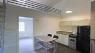 This two-floor space is defined by its striking architectural character, most notably the vaulted ceiling that rises dramatically above the main living and kitchen area, creating an expansive, open atmosphere. A metal railing runs along the upper level, overlooking the space below and reinforcing the vertical scale of the interior.

The kitchen is positioned along one wall and is equipped with white cabinetry, a stainless steel sink, and a gas range, providing a practical and clean cooking area. Dark hardwood flooring grounds the kitchen zone, while a small dining table with two chairs sits nearby, offering a compact and convenient dining setup.

A pendant light hangs from above, providing focused illumination over the dining area and adding a subtle design element to the otherwise minimal aesthetic. The adjoining room, visible through a wide doorway, features light-toned carpet and a window with natural light filtering through, contributing to the overall brightness of the space.

The neutral color palette throughout — with white walls and light finishes — keeps the interior feeling clean and versatile. The combination of high ceilings, open sightlines, and a straightforward layout makes this a functional and visually interesting two-floor space.