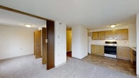 This single-floor unit presents a practical and straightforward layout designed for comfortable everyday living. The main living area features low-pile carpeting and flows openly toward the kitchen and adjoining hallways, creating a connected, easy-to-navigate floor plan. A distinctive dark wood-paneled sliding or folding partition adds a retro architectural character, separating the living space from additional rooms while contributing visual warmth against the otherwise neutral white walls.

The kitchen is outfitted with light honey-toned wood cabinetry providing ample storage, a white electric range, and a refrigerator. Tile flooring in the kitchen zone contrasts with the carpeted living area, clearly delineating functional spaces. The textured ceiling is consistent throughout, adding subtle dimension to the interior.

Hallways branch off toward what appear to be private rooms, hinting at a multi-room configuration suited for various living arrangements. White walls keep the interior bright and neutral throughout.

Perhaps the most notable feature is the expansive urban skyline view visible from the building's elevated position, offering a broad panoramic outlook over a mid-sized city with a mix of residential and commercial architecture, green spaces, and open sky.