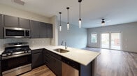 This single-floor space is designed with a contemporary aesthetic, combining functional layout with modern finishes throughout. The kitchen serves as a central highlight, featuring dark charcoal cabinetry with sleek hardware, crisp white countertops, and a herringbone-patterned tile backsplash. A peninsula island with an undermount stainless steel sink and three stylish cylindrical pendant lights overhead provides both additional workspace and a natural division between the kitchen and living area. Stainless steel appliances, including a gas range, microwave, and french door refrigerator, complete the kitchen's polished look. Wood-look flooring runs throughout the main living areas, adding warmth and continuity to the space. The open living area is bright and airy, with natural light streaming in through multiple windows and French doors that lead directly to an outdoor patio area. A ceiling fan provides additional comfort in both the living room and bedroom. The bedroom is generously sized, carpeted for comfort, and features dual windows that invite abundant natural light. The bathroom is finished with marble-style tile in a glass-enclosed walk-in shower, a white countertop vanity with dark cabinetry, and wood-look flooring, maintaining the cohesive modern design found throughout the space.