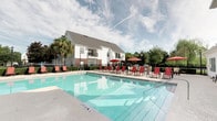 Pooll 360 is a thoughtfully designed outdoor amenity space centered around a generously sized, crystal-clear swimming pool with clearly marked depth indicators. The pool is surrounded by a wide, textured concrete sundeck that provides ample room for relaxation and movement. Rows of red lounge chairs and umbrella-shaded tables are positioned poolside, offering residents comfortable spaces to sunbathe or socialize. A standout feature of the deck is a stone fire pit, ideal for evening gatherings. Adjacent to the pool, a covered outdoor lounge area furnished with plush, red-cushioned wicker sofas and accent pillows creates a cozy retreat beneath the shade of a mature palm tree. A classical-style building with white columns, large windows, and decorative planters with bold blue pots borders one side of the pool, adding an elegant architectural backdrop. Additional amenities such as grilling stations are visible along the perimeter. The entire area is enclosed by a black iron fence, ensuring privacy and safety, while mature palm trees and manicured hedges contribute to a lush, resort-like atmosphere. Signage including pool rules and emergency information reflects a commitment to resident safety and well-organized community management.