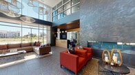 This contemporary communal space makes an immediate impression with its soaring double-height ceilings and expansive floor-to-ceiling windows that flood the interior with natural light and frame views of the surrounding greenery. The flooring throughout is a polished terrazzo, lending a refined, durable finish that anchors the open-plan layout.

At the heart of the space is a reception desk featuring warm wood tones and a welcoming "hello." signage, setting a friendly and professional tone. Adjacent to the entry, a dramatic floor-to-ceiling textured stone fireplace wall serves as a striking focal point, flanked by bold red accent chairs and a casual lounge sofa. Sculptural pendant lighting adds artistic character overhead.

Further into the space, a business center offers individual workstations with built-in desks, providing a quiet area for focused tasks. A teal channel-tufted sofa and bar-height seating around a wood communal table create additional social and collaborative zones. Colorful hexagonal wall art in teal, copper, and charcoal adds a playful, modern touch to the dark feature walls.

A semi-private lounge nook near the entrance features high-back upholstered chairs and a shared media screen, ideal for small group gatherings. A glass-walled conference room with a full boardroom table is also visible, offering a dedicated space for formal meetings. The overall ambiance is modern, vibrant, and thoughtfully designed for both productivity and relaxation.