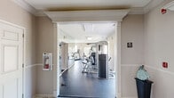 Located at 500 E Centennial Drive in Oak Creek, Wisconsin, this single-story fitness center offers a dedicated exercise room and a welcoming entry hallway. The exercise room is generously sized, featuring durable rubber flooring designed to withstand heavy equipment use. Large windows line multiple walls, flooding the space with natural light and providing pleasant views of the surrounding greenery and outdoor landscape — creating an inviting atmosphere for workouts.

The facility is equipped with a comprehensive selection of cardio and strength-training machines, including treadmills, ellipticals, a stationary bike, and multiple multi-functional cable and weight machines. A wall of floor-to-ceiling mirrors expands the visual footprint of the room while allowing users to monitor their form. Overhead, flush-mounted panel lighting supplements the abundant natural light. A wall-mounted television adds a modern convenience for entertainment during exercise.

The entry hallway features elegant marble-style flooring, providing a clean and polished transition into the fitness area. Neutral, warm-toned walls throughout contribute to a calm and focused environment. The overall design of the space is practical yet refined, balancing functionality with a well-maintained, professional aesthetic suited for everyday fitness use.