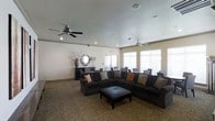 This single-floor lounge space offers a comfortable and well-appointed gathering area designed for relaxation and socializing. The main seating area is anchored by a large, dark sectional sofa accented with decorative throw pillows in warm tones, complemented by a tufted leather ottoman that serves as both a centerpiece and functional surface. The room is finished with a bold leopard-print carpet that adds a distinctive character to the space.

Two ceiling fans are mounted overhead, providing efficient air circulation throughout the generously sized room. Recessed lighting supplements the natural light that pours in through multiple large windows dressed with horizontal blinds, creating a bright and inviting atmosphere.

Along one wall, a dark wood console table is styled with a decorative vase and a sunburst mirror, adding an elegant focal point to the room. Adjacent to the main lounge area, a secondary zone features additional seating with patterned armchairs arranged around small tables, making it well-suited for smaller group conversations or individual use. The neutral wall tones provide a clean backdrop that ties the room's varied design elements together cohesively.