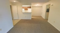 This single-floor unit offers a straightforward and functional layout designed to maximize open living space. The living area is finished with wall-to-wall neutral carpeting that extends throughout, creating a cohesive and comfortable feel underfoot. A half-wall pass-through connects the kitchen to the main living space, promoting an open, airy atmosphere while maintaining a subtle sense of separation between the two areas. The kitchen features warm wood-toned cabinetry, a white refrigerator, and light countertops, providing ample storage and a clean, practical workspace. Ceiling-mounted lighting keeps the kitchen well-lit, while the bright white walls throughout the unit contribute to a clean, neutral aesthetic that serves as an easy canvas for personalization. A doorway visible to the side of the living area leads to additional rooms, suggesting a sensible floor plan with clear circulation. The tile flooring at the kitchen entry transitions smoothly to the carpeted living zone, adding a subtle material contrast. Overall, the space presents a clean, move-in-ready interior with a practical open-plan design suited to everyday living.