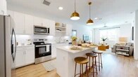 This thoughtfully designed 2-bedroom, 2-bathroom single-story residence offers a clean, modern aesthetic defined by crisp white walls, light wood-tone flooring, and warm gold accents throughout. The open-concept kitchen serves as a central hub, featuring white shaker cabinetry, stainless steel appliances, subway tile backsplash, quartz countertops, and a center island with bar seating illuminated by striking brass pendant lights. The kitchen flows seamlessly into the living area, creating an inviting, connected space filled with natural light from large windows. Both bedrooms are generously proportioned, with the spacious primary bedroom offering abundant room for full-sized furnishings, large windows, a ceiling fan, and access to a well-organized walk-in closet with built-in shelving and hanging rods. Each bathroom is finished with white cabinetry, quartz vanity tops, large mirrors, and wood-look flooring. The primary bathroom features a walk-in shower, while the secondary bathroom includes a soaking tub and shower combination. Two dedicated closets provide ample storage throughout the home. A welcoming entrance and connecting hallway complete the efficient, functional floor plan, making this a comfortable and stylish single-floor residence.
