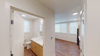 This well-designed single-floor unit offers a clean, modern aesthetic with consistent warm-toned hardwood flooring and crisp white walls throughout. The kitchen is a functional highlight, featuring light wood cabinetry that extends to the ceiling for ample storage, a white quartz countertop, and a full suite of stainless steel appliances including a refrigerator, electric range, over-the-range microwave, and dishwasher. Natural light filters in through a side window, brightening the workspace. Adjacent to the kitchen, a hallway connects the remaining areas of the unit seamlessly. The bedroom is a calm, open space illuminated by roller-shaded windows that allow for adjustable natural light. Recessed ceiling lighting complements the room, and the hardwood flooring continues throughout, maintaining a cohesive look. The bathroom is well-appointed with a light wood vanity topped with white quartz, a large wall mirror with overhead lighting, a toilet, and a separate bathtub and shower area with tiled surrounds. A window provides natural ventilation. Overall, the unit presents a fresh, contemporary design with practical layouts, quality finishes, and a bright, inviting atmosphere.