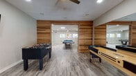 This single-floor game room is thoughtfully designed to provide a dynamic and engaging recreational experience. The space is divided into interconnected areas separated by striking warm-toned wood panel accent walls, which lend a modern yet inviting character to the environment. Durable light gray luxury vinyl plank flooring runs throughout, providing a clean, cohesive foundation that complements the contemporary aesthetic.

The main area is equipped with multiple game tables, including two pool tables, a foosball table, and a shuffleboard table, offering a well-rounded selection of entertainment options. Large floor-to-ceiling mirrors are strategically positioned along the walls, visually expanding the space and enhancing the sense of openness. Wall-mounted flat-screen televisions are present in multiple zones, adding a multimedia component to the recreational experience.

Natural light enters through windows along the exterior walls and glass-paneled doors, keeping the atmosphere bright and welcoming. Ceiling fans with integrated lighting are installed throughout, ensuring comfortable airflow and consistent illumination. Recessed lighting further supplements the overall brightness of the space.

The adjacent room, accessible through the wood-paneled partition, provides additional gaming space and benefits from its own ceiling fan, windows, and mirrored wall panels. Together, the zones create a versatile, well-appointed game room suited for casual socializing and active play alike.