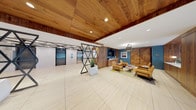 This single-floor commercial lobby space blends contemporary design with warm, natural materials to create a sophisticated and inviting environment. The standout feature is the rich wood-paneled ceiling that extends throughout, adding warmth and texture, complemented by recessed lighting that ensures bright, even illumination across the space.

The entry area welcomes visitors with floor-to-ceiling glass panels and a statement wood-framed door, allowing abundant natural light to flood the interior. Large-format light-toned tile flooring flows seamlessly throughout, enhancing the sense of openness and scale.

A dramatic pair of geometric, lattice-style metal sculptures flanks the elevator bank, serving as bold architectural focal points. The elevators themselves are finished with clean, modern materials and framed by additional wood paneling.

The lounge area is furnished in a mid-century modern style, featuring mustard-yellow accent chairs, a low coffee table, and dark lounge seating arranged around a teal blue feature wall with mounted displays. Wood barn-style doors add rustic character to this zone. A built-in reception or concierge desk anchors one side of the space.

Adjacent to the entrance, a glass-walled office or conference room provides visual transparency while maintaining separation. Potted plants throughout add a touch of greenery. Together, these elements create a professional yet warm and welcoming atmosphere.