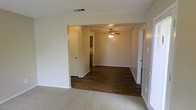 This single-story home in Arlington, Texas offers a straightforward, functional layout with a clean, neutral aesthetic. The interior showcases a combination of plush carpet in the main living area and dark hardwood-style flooring in the adjoining space, creating a clear visual distinction between zones while maintaining a cohesive overall look. The open passageway connecting the two areas enhances the sense of flow throughout the home, making the layout feel open and accessible.

The main living area features light-toned carpet and crisp white walls, while the adjacent room—visible through a wide arched opening—is fitted with a ceiling fan with integrated lighting, providing both comfort and illumination. This secondary space opens further toward what appears to be a kitchen area, hinted at by cabinetry visible in the background.

Multiple interior doors provide access to additional rooms, suggesting a well-organized floor plan. The textured ceiling and neutral wall tones throughout contribute to a simple, understated ambiance. HVAC ventilation is integrated into the ceiling, ensuring year-round comfort. Overall, the space presents a practical and move-in-ready environment suitable for a variety of living arrangements.