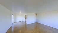 This single-floor space is characterized by its open, airy layout and minimalist design aesthetic. The main living area is notably spacious, with smooth white walls and a clean, flat ceiling that together create a bright and neutral atmosphere. Natural light filters in through a window or sliding door visible along one wall, contributing to the overall sense of openness.

The flooring throughout the main area consists of parquet-style tiles arranged in a block pattern, lending a classic and understated character to the space. A partial dividing wall separates the living area into distinct zones without fully closing off the floor plan, maintaining a sense of flow and connectivity between spaces.

Beyond the dividing wall, a doorway leads to what appears to be a bathroom, identifiable by visible fixtures, while an additional doorway provides access to a separate room or corridor. A baseboard heating unit runs along one wall, indicating a practical heating solution integrated into the architecture.

The overall ambiance of the space is clean, simple, and move-in ready, offering a neutral canvas well-suited to a variety of furniture arrangements and interior design preferences. The uncluttered design allows the architectural details — particularly the parquet flooring and open layout — to stand out as defining features of the space.