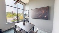 Located at 4825 Ellin Rd in Hyattsville, Maryland, this refined dining room offers a clean, contemporary aesthetic designed to impress. The space is anchored by a sleek marble-top table surrounded by upholstered chairs, set atop a richly textured teal-toned carpet that adds depth and warmth to the neutral palette of crisp white walls. Soaring ceilings amplify the sense of openness, while a striking copper-finished linear pendant light overhead provides both functional illumination and a sophisticated design accent. A wall-mounted flat-screen TV makes the room equally suited for presentations or media viewing. Large floor-to-ceiling windows flood the interior with abundant natural light and offer views of the surrounding greenery and streetscape, creating a pleasant, airy atmosphere. A tall, arched black display cabinet with glass doors houses curated white ceramic vases and books, serving as an eye-catching decorative focal point. A framed art piece adorns the adjacent wall, contributing to the room's polished, gallery-like character. A dark-framed glass door connects the space to the wider facility, maintaining visual continuity while ensuring privacy. Overall, this space strikes a confident balance between functional utility and refined style.