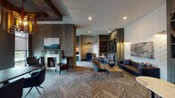 Located in Humble, Texas, this single-floor library lounge is a thoughtfully designed communal space that blends refined aesthetics with functional amenities. The interior is unified by striking herringbone-patterned carpeting that flows throughout, anchoring the various seating arrangements and zones within the space.

The lounge area features an eclectic yet cohesive mix of furnishings, including a tufted navy sofa accented with decorative pillows, plush velvet accent chairs in warm caramel tones, and an abstract-patterned bench seat. A sleek rectangular coffee table serves as a central focal point. A fireplace with a clean white surround adds warmth to one corner, framed by textured herringbone wallcovering and complemented by curated artwork.

A geometric built-in shelving unit finished in muted sage tones displays decorative objects and books, functioning as both a design feature and a room divider. Adjacent to it, a cozy nook with a bistro-style table and chairs provides an intimate spot for casual conversation or individual use.

A dedicated workstation area at the rear accommodates multiple users, outfitted with desks, monitors, and pendant lighting. A service counter with wood-panel cabinetry and decorative sculptural accents rounds out the space. The overall ambiance is sophisticated and inviting, with layered lighting, dimensional wall textures, and curated art enhancing the upscale atmosphere throughout.