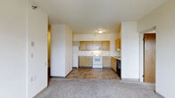 This single-floor space presents a practical and functional layout centered around an open-concept design that connects the living area to the kitchen. The kitchen is equipped with warm-toned wooden cabinetry offering ample upper and lower storage. Essential white appliances are included, featuring a refrigerator, electric range, and a dishwasher, along with an under-cabinet range hood. The countertops provide sufficient prep space, and a sink is positioned along the side wall. The kitchen flooring consists of neutral tile in a classic pattern, providing durability in a high-use area. The adjacent living space transitions seamlessly into carpeted flooring in a muted, neutral tone, adding comfort underfoot. Flush-mounted ceiling lighting illuminates both areas effectively. The walls throughout are finished in a clean, off-white tone, contributing to a bright and open atmosphere. A hallway visible to the side suggests additional rooms beyond the main living and kitchen space. Overall, the layout prioritizes functionality and efficient use of space within a straightforward, no-frills design.
