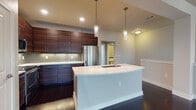 This two-story residence showcases a contemporary design with a thoughtful layout distributed across two levels. The kitchen serves as a standout feature, boasting rich, dark wood grain cabinetry extending to the ceiling, a mosaic tile backsplash, and a full suite of stainless steel appliances including a refrigerator, range, and built-in microwave. A large curved island with a crisp white countertop and undermount sink anchors the space, illuminated by sleek pendant lights that hang from the elevated ceiling. Dark hardwood flooring flows through the kitchen and main living area, adding warmth and contrast against the neutral walls.

The living area offers an open, versatile space with the same dark hardwood floors and clean white walls. Bedrooms are finished with plush carpet and feature windows with blinds that allow natural light to filter in. Bathrooms are well-appointed throughout; one full bath includes a tiled tub-shower combination, while a second features a dual-sink vanity with white countertops, dark wood cabinetry, and large framed mirrors. A powder room with tile flooring provides convenient half-bath facilities. The overall aesthetic is modern and cohesive.