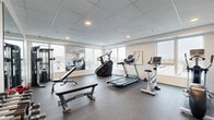 Located at 1530 First Ave N in Moorhead, Minnesota, this fitness center is a compact yet well-equipped exercise facility housed within a single room. The space features a drop-panel ceiling with recessed lighting and durable rubber flooring designed to withstand heavy equipment use. Large windows line the walls, allowing abundant natural light to fill the room while offering views of the surrounding neighborhood. Full-length wall mirrors further enhance the sense of openness and allow users to monitor their form during workouts.

The exercise room is outfitted with a diverse selection of cardio equipment, including treadmills, a stair climber, a stationary bike, and an elliptical machine. A cable weight machine and adjustable bench provide strength training options, while a three-tier dumbbell rack stocked with a range of hex weights supports free weight workouts. Framed wall art featuring a nature-inspired landscape adds a subtle decorative touch to the otherwise functional space.

A glass-paneled entry door provides access to the fitness center, and the neutral gray walls contribute to a clean, modern aesthetic throughout. The layout is thoughtfully organized to accommodate both cardio and strength training within an efficient footprint, making this fitness center a practical and well-rounded amenity.