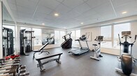 Located at 1530 First Ave N in Moorhead, Minnesota, this fitness center is a compact yet well-equipped exercise facility housed within a single room. The space features a drop-panel ceiling with recessed lighting and durable rubber flooring designed to withstand heavy equipment use. Large windows line the walls, allowing abundant natural light to fill the room while offering views of the surrounding neighborhood. Full-length wall mirrors further enhance the sense of openness and allow users to monitor their form during workouts.

The exercise room is outfitted with a diverse selection of cardio equipment, including treadmills, a stair climber, a stationary bike, and an elliptical machine. A cable weight machine and adjustable bench provide strength training options, while a three-tier dumbbell rack stocked with a range of hex weights supports free weight workouts. Framed wall art featuring a nature-inspired landscape adds a subtle decorative touch to the otherwise functional space.

A glass-paneled entry door provides access to the fitness center, and the neutral gray walls contribute to a clean, modern aesthetic throughout. The layout is thoughtfully organized to accommodate both cardio and strength training within an efficient footprint, making this fitness center a practical and well-rounded amenity.