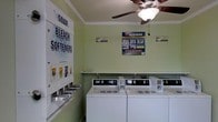 This single-floor shared laundry facility is designed with functionality and convenience in mind. The room is finished with light green walls and crown molding, giving it a clean, welcoming atmosphere. A ceiling fan with an integrated light fixture provides both ventilation and illumination, keeping the space comfortable during use. The room is equipped with multiple top-load commercial washing machines lined along the wall, offering ample capacity for residents or guests. A dedicated laundry supply dispenser panel is mounted on the wall, offering easy access to essentials such as soap, bleach, and fabric softener — a thoughtful amenity for those who may not have supplies on hand. Informational signage is posted throughout the room, providing users with guidance on operating the machines and care instructions. A small shelf is also available, providing a convenient surface for placing personal items during laundry cycles. The overall layout is efficient and organized, making the space easy to navigate. The room reflects a practical design approach, prioritizing user convenience through clearly labeled dispensers, well-maintained equipment, and clear posted instructions.