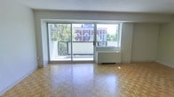 This single-floor space is characterized by its open layout and abundant natural light. The main living area features classic parquet wood flooring arranged in a geometric pattern, lending warmth and texture underfoot. Large sliding glass doors span much of the exterior wall, seamlessly connecting the interior to a private balcony enclosed by a sleek metal railing. This outdoor space offers pleasant views of mature trees and a notable red-brick heritage-style building, creating an attractive urban backdrop. The sliding doors allow generous amounts of natural light to fill the room throughout the day. A built-in white radiator unit is positioned along the exterior wall, providing in-unit heating. The interior walls are finished in a neutral white tone, complementing the light wood flooring and contributing to an airy, clean aesthetic. The overall ambiance is bright and functional, with the combination of parquet floors, large glazed openings, and balcony access creating a comfortable and visually appealing living environment well-suited to urban living.