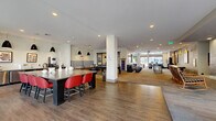Located at 1550 Raleigh St in Denver's vibrant West Colfax neighborhood, this single-floor amenity space delivers a dynamic and modern environment designed for both social interaction and productive work. The layout flows across several distinct yet interconnected zones, all unified by warm hardwood-style flooring, recessed lighting, and large floor-to-ceiling windows that invite abundant natural light and outdoor views.

The main clubroom is a spacious, open-plan gathering area featuring a bold bar-height communal table with red chairs, pendant lighting, and a striped lounge booth. A billiards table anchors the game zone nearby, adding a recreational focal point. Adjacent to this is a comfortable lounge area furnished with plush sofas, accent chairs, a wall-mounted TV, and curated artwork, complemented by a tile-surround fireplace and direct access to an outdoor area.

The lobby and mail center area showcases a striking teal accent wall, a large built-in mailbox bank, terrazzo flooring, and an oversized island counter beneath geometric pendant lights. Decorative textured panels with colorful accents add visual interest throughout.

A dedicated conference room offers a large dark wood table, rolling chairs, a wall-mounted display, and views toward the street entrance — ideal for meetings or private gatherings. The overall ambiance is contemporary, welcoming, and thoughtfully designed for community living.
