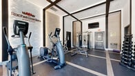 The fitness center at Rachel Gardens Montville in Pine Brook, NJ, is a thoughtfully designed exercise space that balances modern aesthetics with comprehensive workout functionality. The room features soaring ceilings accented by bold dark structural beams arranged in a geometric grid pattern, lending the space a striking architectural character. A series of arched windows along the exterior walls flood the interior with abundant natural light, offering outdoor views and an open, airy atmosphere.

The flooring consists of durable dark rubber gym tiles with light-toned inlay lines that mirror the ceiling grid above, creating a cohesive and polished visual design. Full-length mirrors are strategically positioned along the walls to amplify the sense of space and support proper exercise form.

The facility is comprehensively equipped for a full-body workout. Cardio options include multiple treadmills, elliptical trainers, a stationary cycle, and an Assault fitness rower. Strength training is supported by a complete dumbbell rack, a weight bench, and a multi-function cable machine. A wall-mounted flat-screen television adds entertainment convenience. The Rachel Gardens Montville branded feature wall — a sculpted, textured panel with warm accent lighting — serves as a distinctive focal point, reinforcing the upscale identity of the community.