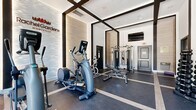 The fitness center at Rachel Gardens Montville in Pine Brook, NJ, is a thoughtfully designed exercise space that balances modern aesthetics with comprehensive workout functionality. The room features soaring ceilings accented by bold dark structural beams arranged in a geometric grid pattern, lending the space a striking architectural character. A series of arched windows along the exterior walls flood the interior with abundant natural light, offering outdoor views and an open, airy atmosphere.

The flooring consists of durable dark rubber gym tiles with light-toned inlay lines that mirror the ceiling grid above, creating a cohesive and polished visual design. Full-length mirrors are strategically positioned along the walls to amplify the sense of space and support proper exercise form.

The facility is comprehensively equipped for a full-body workout. Cardio options include multiple treadmills, elliptical trainers, a stationary cycle, and an Assault fitness rower. Strength training is supported by a complete dumbbell rack, a weight bench, and a multi-function cable machine. A wall-mounted flat-screen television adds entertainment convenience. The Rachel Gardens Montville branded feature wall — a sculpted, textured panel with warm accent lighting — serves as a distinctive focal point, reinforcing the upscale identity of the community.