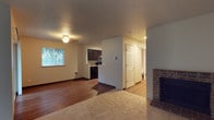 This two-floor space presents an open and functional main level that seamlessly connects the living area, kitchen, and hallway. The living room is anchored by a brick fireplace on one side, adding warmth and a classic architectural detail to the space. Soft carpet covers the living area, while rich hardwood flooring extends through the adjacent dining and kitchen zones, creating a natural visual distinction between areas. A window on the front wall allows natural light to filter into the living space, brightening the neutral, cream-colored walls. The kitchen features dark wood cabinetry paired with black appliances, including a range and over-the-range microwave, offering a modern contrast to the lighter tones of the surrounding space. A textured popcorn ceiling is consistent throughout the main level, complemented by flush-mount and recessed lighting. A central hallway with a six-panel door leads toward additional rooms, with light-toned flooring continuing through that corridor. A wall-mounted thermostat and floor-level heating vent are also visible, indicating climate control throughout. The layout is practical and well-connected, with the second floor offering additional living space above.