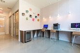 Located in Atlanta, GA (30316), this contemporary single-floor coworking and office space is thoughtfully designed to support a range of professional needs. The layout is anchored by a bright, polished corridor lined with floor-to-ceiling glass-paneled doors and walls, creating a sense of openness and transparency throughout. Natural light pours in through large, expansive windows that frame views of the surrounding urban streetscape, enhancing the airy, modern atmosphere.

Enclosed conference rooms feature sliding glass doors, wooden tables, and vibrant red ergonomic chairs set atop colorful geometric-patterned carpet tiles — a striking contrast to the clean white walls and smooth resin flooring found in the common areas. Smaller private rooms, also glass-fronted and individually keyed, offer focused workspaces suitable for individual use or small meetings.

A communal workstation area provides built-in countertop desks, computer terminals, and a printer, bathed in natural light from floor-to-ceiling windows. A beverage refrigerator is conveniently integrated beneath the counter. A playful accent wall adorned with a colorful collection of vintage-style telephones adds a creative, character-driven touch to the space. The overall design balances professionalism with personality, making it a functional and visually engaging environment for modern work.