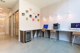 Located in Atlanta, GA (30316), this contemporary single-floor coworking and office space is thoughtfully designed to support a range of professional needs. The layout is anchored by a bright, polished corridor lined with floor-to-ceiling glass-paneled doors and walls, creating a sense of openness and transparency throughout. Natural light pours in through large, expansive windows that frame views of the surrounding urban streetscape, enhancing the airy, modern atmosphere.

Enclosed conference rooms feature sliding glass doors, wooden tables, and vibrant red ergonomic chairs set atop colorful geometric-patterned carpet tiles — a striking contrast to the clean white walls and smooth resin flooring found in the common areas. Smaller private rooms, also glass-fronted and individually keyed, offer focused workspaces suitable for individual use or small meetings.

A communal workstation area provides built-in countertop desks, computer terminals, and a printer, bathed in natural light from floor-to-ceiling windows. A beverage refrigerator is conveniently integrated beneath the counter. A playful accent wall adorned with a colorful collection of vintage-style telephones adds a creative, character-driven touch to the space. The overall design balances professionalism with personality, making it a functional and visually engaging environment for modern work.