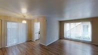 This single-floor space offers a clean, functional layout with a welcoming interior design. The living area is finished with rich wood-look laminate flooring that extends throughout, providing a warm and cohesive aesthetic. Neutral-toned walls in soft beige and gray create a versatile backdrop suited to a variety of furnishing styles.

A large window fitted with vertical blinds anchors the main living area, allowing ample natural light to fill the room while offering privacy control. The textured ceiling adds a classic architectural detail found throughout the space.

Adjacent to the living area, a designated dining space is elevated by an elegant chandelier that adds a decorative focal point and warm ambient lighting. This area flows naturally toward the kitchen, which is visible through an open doorway and features white cabinetry and appliances, contributing to a bright and tidy culinary space.

The overall layout is open and efficiently organized, making the most of the available floor plan. The combination of natural light, hardwood-style flooring, and thoughtful lighting fixtures gives the space a comfortable and inviting atmosphere that is both practical and aesthetically appealing.