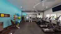This fitness center is thoughtfully designed to accommodate a wide range of workout styles and fitness goals. The space features a clean, modern aesthetic with a neutral gray and vibrant teal color scheme, durable diamond-plate rubber flooring, and recessed lighting complemented by dual ceiling fans that ensure comfortable air circulation throughout.

The cardio section is well-stocked with multiple treadmills, elliptical trainers, and a stationary spin bike, all arranged to face a full wall of floor-to-ceiling mirrors that visually expand the space and allow users to monitor their form. Mounted flat-screen TVs provide entertainment during workouts. Large windows along one wall invite natural light and offer views of the surrounding outdoor area.

A dedicated TRX Training Zone is clearly marked along the vibrant teal accent wall, and includes suspension training straps, workout instruction charts, a weight bench, and a rack stocked with medicine balls, resistance bands, and exercise mats. Additional strength training equipment and a cable machine round out the functional fitness options.

A motivational wall decal reading "The Body Achieves What the Mind Believes" adds an inspiring focal point to the space. A hand sanitizer station and informational signage reflect attention to cleanliness and user convenience. The fitness center is accessible via a dedicated corridor, making it easy to locate and use.