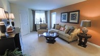 This single-floor residence offers a comfortable and well-appointed living space designed with both style and functionality in mind. The living room serves as the heart of the home, showcasing plush wall-to-wall carpeting in a warm neutral tone that creates a cozy, welcoming atmosphere throughout. A large bay window floods the space with natural light, framed by elegant floor-length drapes that add a refined touch to the room. The warm terracotta accent wall provides a striking focal point, complemented by a curated gallery of framed artwork that adds visual depth and character. Seating is generously arranged, with a plush sofa adorned with decorative accent pillows positioned alongside a pair of classic armchairs, all centered around a rich, dark-wood round coffee table. A secondary window with a door panel offers additional light and access, enhancing the open feel of the space. The overall ambiance is one of understated elegance — a harmonious blend of warm earth tones, tasteful décor, and practical layout that makes the living area both aesthetically pleasing and highly livable. The single-level design ensures seamless flow and easy accessibility throughout the residence.