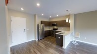 This single-floor space showcases a contemporary open-concept layout, with the kitchen serving as a central highlight. The kitchen is outfitted with a full suite of stainless steel appliances, including a French door refrigerator, electric range with microwave above, and an integrated sink set within a clean white countertop. Dark espresso-toned cabinetry lines the walls, providing ample storage while adding a bold contrast to the light-colored walls and countertops. A peninsula with a white waterfall edge extends into the living area, creating a functional breakfast bar space ideal for casual dining or entertaining. Pendant lighting hangs above the peninsula, offering focused illumination and a modern aesthetic touch. Recessed lighting throughout the ceiling ensures the space is well-lit. The flooring features medium-toned hardwood-style planks that run continuously, lending warmth and visual cohesion to the open layout. The overall ambiance is clean, modern, and functional, with a neutral color palette of whites, grays, and dark browns that creates a sophisticated yet inviting atmosphere. A keypad-secured entry door is visible, suggesting a secure and contemporary building environment.