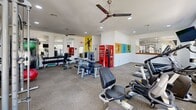 This fitness center in Phoenix, AZ offers a well-equipped, single-floor exercise room designed to support a comprehensive workout experience. The space is finished with neutral carpet flooring and bright white walls, creating a clean and energizing atmosphere. Large windows along one wall flood the room with natural light and provide views of the surrounding outdoor area, including palm trees and what appears to be a pool area, adding a refreshing Arizona ambiance to every workout.

The exercise room is outfitted with a strong selection of Matrix-brand cardio equipment, including multiple elliptical trainers, stationary bikes, and treadmills, all equipped with individual monitors for entertainment during workouts. A cable machine and selectorized strength stations anchor the strength training section, complemented by a dumbbell rack and stability balls for versatile training options. Ceiling fans and recessed lighting keep the environment comfortable, while wall-mounted televisions add convenience throughout.

Vibrant, athletic-themed wall murals and colorful artwork add personality and motivation to the space. A Coca-Cola vending machine provides convenient refreshment access. The fitness center is well-organized, maximizing its footprint to accommodate a variety of fitness needs within a compact, functional layout ideal for residents or guests seeking a reliable on-site workout facility.