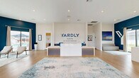 Located at 200 Villas Dr in Lavon, TX, this single-story welcome and leasing center for Yardly Elevon by Taylor Morrison offers a thoughtfully designed, open-concept space that makes a strong first impression. The interior is defined by light hardwood flooring, soaring ceilings, and a crisp white-and-navy color palette that conveys a clean, contemporary aesthetic throughout.

Upon entry through large glass-panel doors, visitors are greeted by a prominently branded reception desk, backlit with subtle accent lighting and anchored by a bold "Yardly Elevon by Taylor Morrison" wall logo. The main area flows openly, featuring multiple distinct zones including a formal reception counter, a comfortable lounge seating area with a channel-tufted sofa and accent chairs, and a casual workstation nook with gray cabinetry and open shelving.

A striking deep-blue accent wall runs through the space, adding depth and visual contrast against the otherwise neutral tones. Large windows and sliding glass doors along the rear wall flood the interior with natural light and offer views of the surrounding community and outdoor areas. A media display station with herringbone wood-panel detailing and a mounted TV serves as an engaging feature wall. Recessed lighting complements the natural light throughout, creating a bright, professional, and inviting environment ideal for welcoming prospective residents.