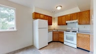 This single-floor space is designed with practicality and comfort in mind. The kitchen serves as a central feature, showcasing warm honey-toned oak cabinetry that extends to the upper walls, providing generous storage throughout. The layout follows a efficient corner configuration, making smart use of the available space. White appliances, including a full-size refrigerator, a gas range with oven, and a dishwasher, are seamlessly integrated into the design. A corner sink with a single pendant light above adds a functional focal point, while the under-cabinet range hood above the stove ensures proper ventilation. The kitchen floor features light-toned tile, which transitions to a neutral carpeted area nearby, delineating the spaces naturally. Flush-mount ceiling lighting keeps the kitchen well-illuminated, and light-colored walls throughout contribute to an open, airy feel. The overall aesthetic is clean and straightforward, offering a move-in-ready environment with all essential kitchen amenities thoughtfully arranged for day-to-day convenience.