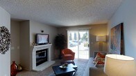 This single-floor space centers around a well-appointed living room that balances comfort and functionality. A white brick fireplace serves as a focal point along one wall, topped with a flat-screen television for a practical yet classic arrangement. The textured popcorn ceiling adds a characteristic mid-century touch to the room's architecture.

Natural light fills the space through a large sliding glass door fitted with vertical blinds, offering views of mature trees and greenery outside. The door provides both an airy connection to the outdoors and easy access to the exterior. Neutral gray walls create a versatile backdrop, complemented by plush carpeting underfoot.

The living area is furnished with a mix of seating options, including a warm terracotta-toned armchair and a gray sofa accented with patterned throw pillows in orange and floral motifs. A dark coffee table anchors the seating arrangement, while a tall floor lamp in the corner contributes warm ambient lighting. A large indoor plant adds a natural, organic element to the space, enhancing its inviting atmosphere. Overall, the room presents a relaxed and livable environment with several characterful details.