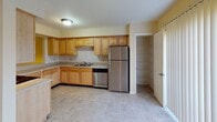 This three-floor residence at Chesterfield Village Apartments offers a well-designed 2-bedroom, 2.5-bathroom layout with generous living space spread across multiple levels. The main living area is immediately impressive, featuring soaring double-height ceilings with an open upper-level overlook, plush carpeting, and a classic fireplace with a white mantel that serves as the room's focal point. Natural light filters in through a sliding glass door that opens to a private balcony with outdoor views. The kitchen is efficiently laid out with warm maple cabinetry, stainless steel appliances including a refrigerator and dishwasher, a built-in pantry, and a convenient pass-through window that connects directly to the living area. The bathrooms are clean and functional, with the full bath featuring tile flooring, a tub-shower combination, and a vanity with ample counter space. Additional bathrooms mirror this clean, neutral aesthetic. The lower level includes a utility or storage room with exposed ductwork and concrete flooring, offering practical flex space. Throughout the residence, a soft, neutral color palette of warm creams and beiges creates a cohesive and inviting atmosphere. Carpeted bedrooms provide comfort, while tiled areas in the kitchen and bathrooms add durability and ease of maintenance.