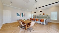 Located at 2150 NW 21st St in Ocala, Florida, this single-floor community gathering space combines modern design with functional versatility. The open-concept layout is anchored by a striking herringbone-patterned light wood floor that runs throughout, lending warmth and visual interest to the entire room.

The fully equipped kitchen is a standout feature, showcasing sleek grey cabinetry, a white quartz island with bar-height leather seating, open floating shelves, a green glass tile backsplash, stainless steel appliances including a French door refrigerator, and a utility sink. Elegant black and gold pendant lights hang above the island, adding a refined touch to the space.

Adjacent to the kitchen is a spacious conference and dining area centered around a solid wood farmhouse-style table surrounded by tan leather swivel chairs on casters — ideal for meetings, gatherings, or collaborative work sessions. Crown molding accents the walls throughout, and recessed lighting provides bright, even illumination.

A defining architectural element is the full-glass panel garage-style door that opens to the outdoors, flooding the space with natural light and creating a seamless indoor-outdoor connection. Contemporary artwork on the walls completes the modern, professional aesthetic, making this a welcoming and highly functional shared space.