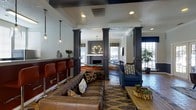This single-story space functions as a sophisticated communal lounge, thoughtfully designed to accommodate both social gatherings and relaxed leisure. The layout is anchored by bold dark-painted structural columns that define distinct zones throughout the open floor plan, creating an organized yet connected atmosphere.

A bar area along one side features rich red-toned bar stools and pendant lighting with glass globe fixtures, lending an inviting, pub-like warmth to the space. Adjacent to this, a comfortable seating area centers around a tufted brown leather sofa and a patterned accent chair in deep navy, arranged over a layered area rug atop herringbone-patterned tile flooring.

Further into the space, a billiards table serves as a focal recreational feature, positioned beneath a decorative chandelier near a brick fireplace that adds a classic, cozy element to the room. Large windows along the perimeter allow natural light to fill the interior, with views of greenery outside enhancing the overall ambiance.

The design palette blends dark architectural accents, warm leather furnishings, and eclectic decorative textiles, resulting in a space that feels both polished and approachable.