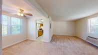 This single-floor unit is laid out with a clear separation between living and functional spaces. The living area is generously sized, finished with neutral wall-to-wall carpeting and crisp white walls. A ceiling fan is installed overhead, and a wall-mounted HVAC unit beneath a double window provides climate control while allowing natural light to filter in. An open rectangular passageway connects the living area directly to the kitchen, creating a practical flow between spaces.

The kitchen is efficiently designed, featuring warm honey-toned oak cabinetry both above and below the countertops, a double-basin sink, laminate countertops, a gas range with an overhead exhaust hood, and a full-size refrigerator. A window above the sink brings in additional natural light. Tile flooring runs throughout the kitchen, contrasting with the carpeted areas elsewhere in the unit.

The bedroom is a quiet, carpeted room with a window fitted with blinds, providing outdoor views and natural light. The electrical panel is accessible within the living space.

The bathroom features classic full-height wall tile in a neutral palette, a soaking tub with shower, a wood-vanity with sink, a framed mirror, and mosaic tile flooring. A window keeps the space naturally lit. The entry foyer transitions from tile to carpet, providing a welcoming introduction to the unit.