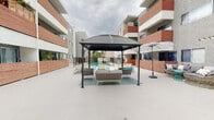 This outdoor amenity space is designed around a central courtyard layout, enclosed by multi-story residential buildings featuring a modern combination of stucco exteriors, brick accent panels, and bold teal architectural highlights. The courtyard is finished with a smooth, light-colored concrete surface that creates a clean, open atmosphere throughout.

A standout feature is the large, covered gazebo fitted with a dark polycarbonate roof, beneath which a generous wicker lounge seating arrangement with teal accent cushions provides a comfortable gathering area. Additional seating options are available across the courtyard, including rounded pod-style chairs, a coordinating sofa set with a patterned area rug, and sleek black lattice chairs positioned near a bowl-shaped fire pit — ideal for relaxed outdoor evenings.

The pool area is secured behind wrought iron fencing with gated access. The swimming pool itself is spacious and rectangular, with vivid turquoise water and brick-edged coping, framed on either side by the surrounding building facades and greenery in the background. Umbrella stands and lounge chairs provide poolside comfort.

The overall ambiance is modern and resort-inspired, balancing functional outdoor living with a cohesive, contemporary aesthetic suited for community recreation and relaxation.