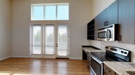 Located at 704 15th Street in Durham, NC, this single-floor studio offers a thoughtfully designed layout with notably high ceilings that enhance the sense of openness throughout. The main living area is a standout feature, showcasing dramatic ceiling height and a set of French doors flanked by large transom windows above, flooding the space with abundant natural light and providing views of surrounding greenery. Light-toned hardwood-style flooring runs through the main living and kitchen areas, adding warmth and continuity.

The kitchen is well-equipped with dark wood-toned cabinetry, open shelving, granite-style countertops, and a full suite of stainless steel appliances including an electric range, over-the-range microwave, dishwasher, and refrigerator. A tiled backsplash and undermount sink complete the functional yet modern kitchen design.

A separate sleeping area features plush carpeting and a clerestory window that brings in additional natural light while maintaining privacy. The bathroom is finished with a granite-topped vanity, a bold framed mirror, oil-rubbed bronze fixtures, and a tub-shower combination with tile surround, delivering a polished and cohesive aesthetic throughout the space.