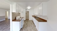 This single-floor unit offers a clean, modern aesthetic throughout, characterized by wood-style plank flooring, textured ceilings, and a neutral color palette of whites and warm grays that creates a bright, cohesive atmosphere.

The galley-style kitchen is efficiently designed with ample white cabinetry on both sides, dark granite-style countertops, a stainless steel sink, a white electric range, and a decorative mosaic tile backsplash. The kitchen opens directly into a comfortable dining or living area outfitted with a ceiling fan and natural light from a window.

The main living area is generously proportioned, featuring sliding glass doors that lead to an outdoor patio, allowing abundant natural light and seamless indoor-outdoor flow. A second window further brightens the space, which carries the same warm wood-style flooring found throughout.

The bedrooms are well-sized, each finished with the same flooring and neutral tones. Several rooms feature walk-in closets, providing ample storage. The two-tone gray and white accent walls add subtle visual interest to the sleeping areas.

The bathroom is cleanly finished in white, featuring a vanity with a white countertop, a wall mirror with overhead lighting, and a separate water closet with a shower-tub combination and a built-in corner shelf.