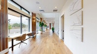 Located at 9875 W 58th Ave in Arvada, Colorado, this expansive single-floor amenity space offers a generously scaled, thoughtfully designed environment ideal for gathering and community engagement. The interior is anchored by wide-plank hardwood flooring that flows throughout, complemented by crisp white walls, exposed wood ceiling beams, and warm walnut slatted accent panels that frame the entryway and key focal points.

The grand entry corridor welcomes visitors with floor-to-ceiling windows, tall potted greenery, and a striking pair of wood-clad columns flanking glass double doors. Natural light pours in from oversized windows along multiple walls, creating a bright and airy atmosphere throughout.

The main lounge area features exposed wood beams overhead, a fireplace with built-in shelving, a wall-mounted display, and plush seating arrangements centered around circular pendant lighting. Adjacent to the lounge, a fully equipped kitchen bar with wood cabinetry, tile backsplash, and counter seating provides a social hub for residents.

A distinct dining nook showcases terracotta-toned wainscoting, arched doorways, banquette seating with marble-topped tables, and decorative wall sconces, lending a boutique café ambiance. Artistic wall sculptures, curated greenery, and cohesive design details tie each area together into a polished, welcoming whole.