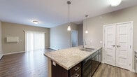 This 2-bedroom, 2-bathroom single-floor unit offers a functional and comfortable layout with a cohesive neutral color palette throughout. The open-concept kitchen and living area serves as the heart of the home, anchored by a large granite-topped island with a double sink and pendant lighting overhead. Dark espresso cabinetry contrasts with the light stone countertops, while black appliances — including a range, microwave, and refrigerator — complete the kitchen. Rich hardwood-style flooring extends through the kitchen and living area, where a sliding glass door fitted with vertical blinds allows natural light to fill the space and provides outdoor access. Both bedrooms feature soft carpeting, neutral walls, and baseboard heating. The primary bedroom is generously sized and includes a walk-in closet with wire shelving for organized storage. The two bathrooms are finished consistently, each featuring granite-topped dark vanities, tub-shower combos, and light tile flooring. A dedicated laundry room houses a full-size side-by-side washer and dryer with overhead shelving. The overall design is clean and practical, offering a well-rounded living experience.