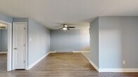 This single-floor space offers a clean, functional layout with a cohesive design throughout. The main living area is open and airy, finished with light wood-look laminate flooring and soft blue-gray walls that create a calm, neutral atmosphere. A ceiling fan with integrated lighting provides both comfort and illumination, while a baseboard heating unit ensures year-round comfort. A wall-mounted air conditioning unit adds additional climate control. Sliding glass doors at the far end of the living space invite natural light and provide direct access to an outdoor area, enhancing the sense of openness.

The kitchen is positioned adjacent to the living area and features rich, dark wood cabinetry paired with light countertops and a decorative mosaic tile backsplash in mixed gray and white tones. Black appliances, including a range and refrigerator, complement the cabinetry, and a window above the sink brings in natural light.

The bedroom is a well-proportioned, private room with the same wood-look flooring, soft blue-gray walls, a ceiling fan, a window with blinds, and baseboard heating. The bathroom is finished with large white wall tiles surrounding a soaking tub and shower combination, gray floor tiles, and a mirrored medicine cabinet, presenting a clean, utilitarian aesthetic throughout.