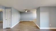 This single-floor space offers a clean, functional layout with a cohesive design throughout. The main living area is open and airy, finished with light wood-look laminate flooring and soft blue-gray walls that create a calm, neutral atmosphere. A ceiling fan with integrated lighting provides both comfort and illumination, while a baseboard heating unit ensures year-round comfort. A wall-mounted air conditioning unit adds additional climate control. Sliding glass doors at the far end of the living space invite natural light and provide direct access to an outdoor area, enhancing the sense of openness.

The kitchen is positioned adjacent to the living area and features rich, dark wood cabinetry paired with light countertops and a decorative mosaic tile backsplash in mixed gray and white tones. Black appliances, including a range and refrigerator, complement the cabinetry, and a window above the sink brings in natural light.

The bedroom is a well-proportioned, private room with the same wood-look flooring, soft blue-gray walls, a ceiling fan, a window with blinds, and baseboard heating. The bathroom is finished with large white wall tiles surrounding a soaking tub and shower combination, gray floor tiles, and a mirrored medicine cabinet, presenting a clean, utilitarian aesthetic throughout.