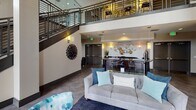 This elegantly designed communal space makes a striking first impression with its dramatic double-height ceilings and open, airy atmosphere. The main lounge area serves as the heart of the space, anchored by a tiled fireplace with a mounted flat-screen television above, flanked by large windows that frame views of the outdoors and flood the room with natural light. The seating area is thoughtfully curated with a plush gray sofa, teal and blue accent pillows, and a geometric-patterned area rug, all arranged around a sleek glass coffee table.

A bold staircase with dark metal railings rises to an upper mezzanine level, which overlooks the lounge below and adds to the sense of vertical grandeur. The space is adorned with distinctive wall art, including a sculptural sunburst installation and abstract canvas paintings, lending it a refined, contemporary character. Pendant and sconce lighting complement the warm, inviting ambiance throughout.

Adjacent to the main lounge, a reception counter with a dark granite surface offers a functional workspace. Nearby, a dedicated fitness center is visible through mirrored glass, equipped with cardio machines. Large-format tile flooring runs consistently throughout, tying the various functional areas together into a cohesive, well-appointed amenity space.